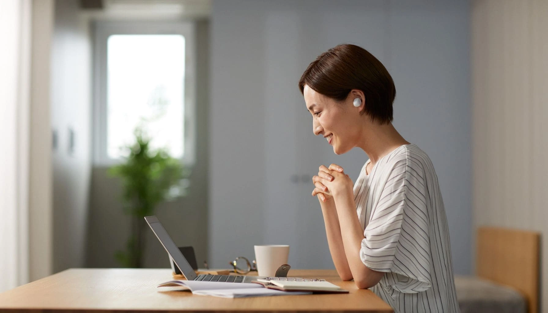 A smiling woman wearing wireless earbuds and looking at a laptop in a bright environment, representing the use of voice technology and AI to enhance patient care and healthcare communication.